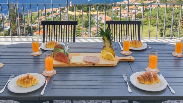 A table set for breakfast with croissants, watermelon, pineapple, and orange juice, overlooking a coastal cityscape. - Bay Bliss by Atlantic Holiday (Funchal)