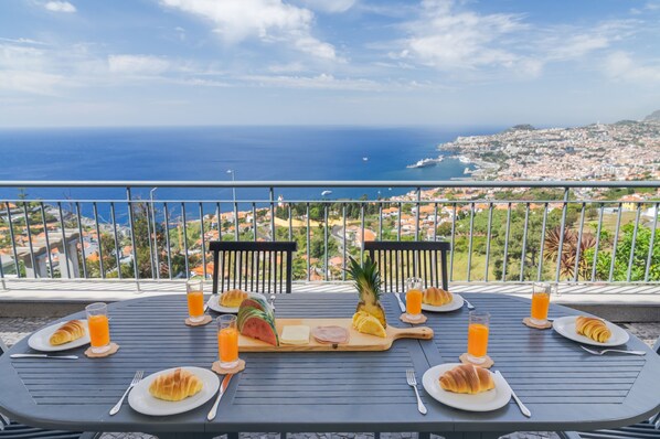 Una mesa dispuesta para el desayuno con croissants, fruta y jugo de naranja, con vistas a una ciudad costera.