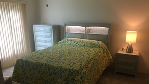 Un dormitorio con una cama, una mesita de noche con lámpara, un aparador y una ventana con persianas.