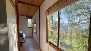 A corridor with tiled flooring, wooden ceiling beams, and large windows offering a view of greenery outside.