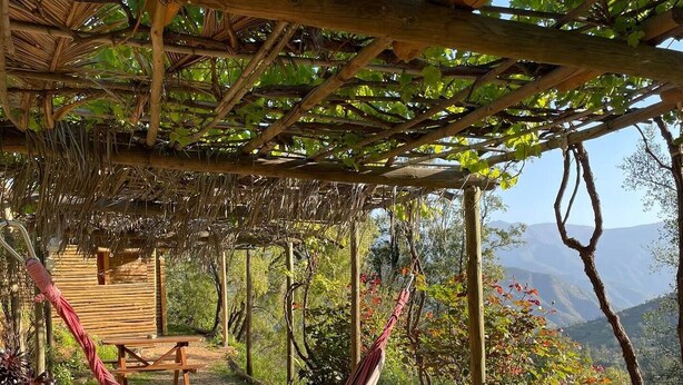 A hammock under a wooden pergola with a thatched roof, a picnic table, and a view of a mountainous landscape.