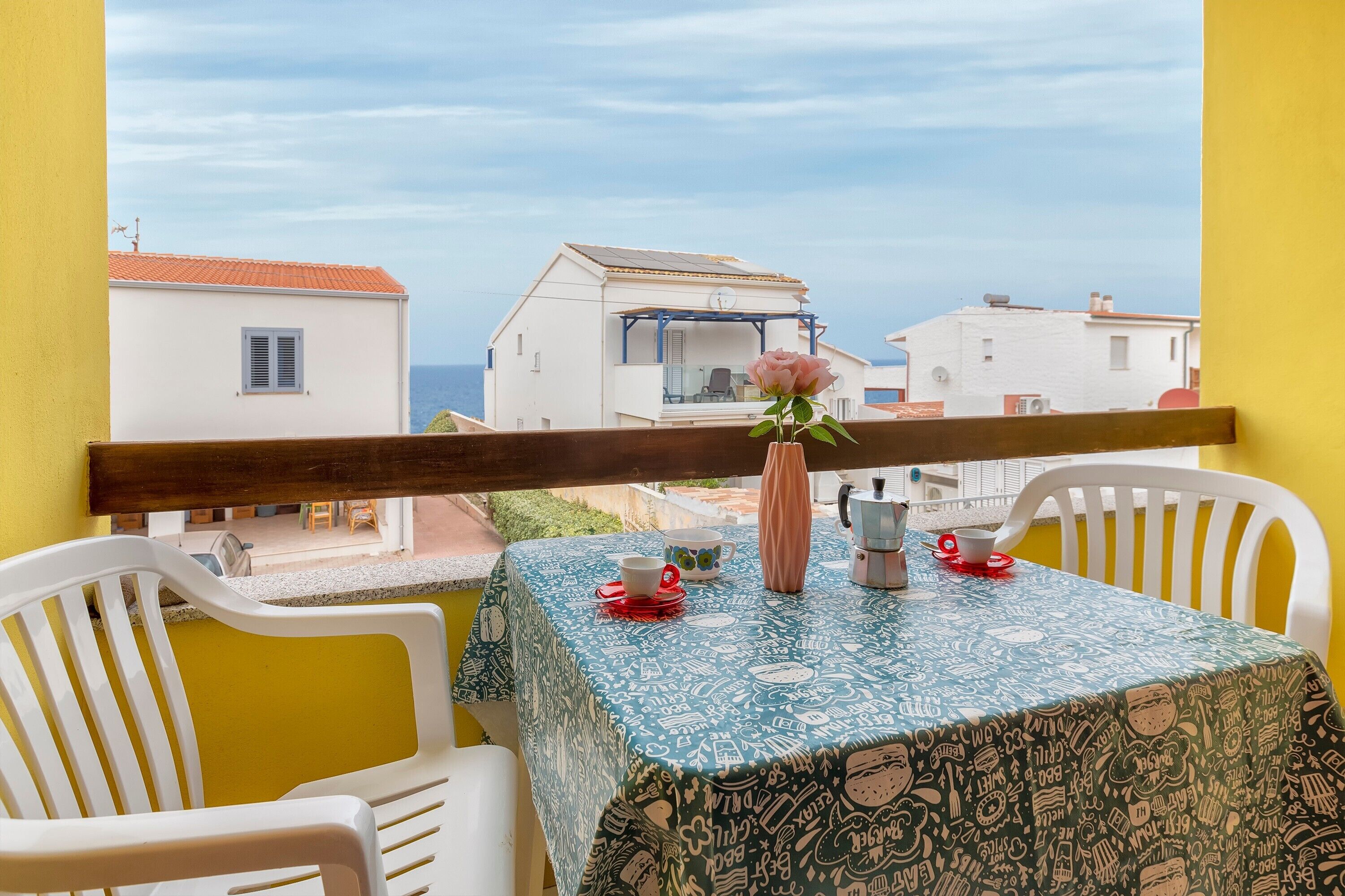 Varanda com mesa posta para dois, vaso com uma flor e vista para edifícios brancos e o mar.