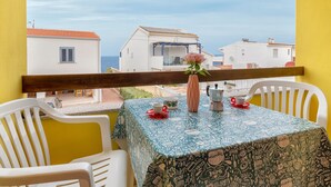 Un balcón con una mesa lista para dos, un jarrón con una sola flor y vistas a edificios blancos y al mar.