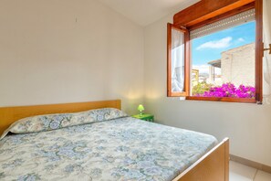 Una camera da letto con un letto, una finestra con tende, un comodino con una lampada e vista su edifici e fiori all'esterno.