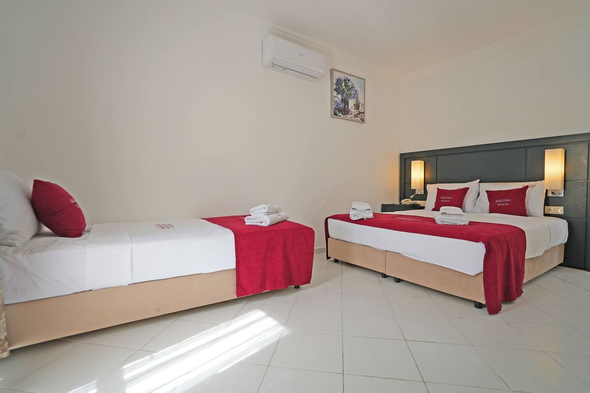 A hotel room with two single beds, each with a red blanket and white pillows, a nightstand, and a wall-mounted air conditioner.