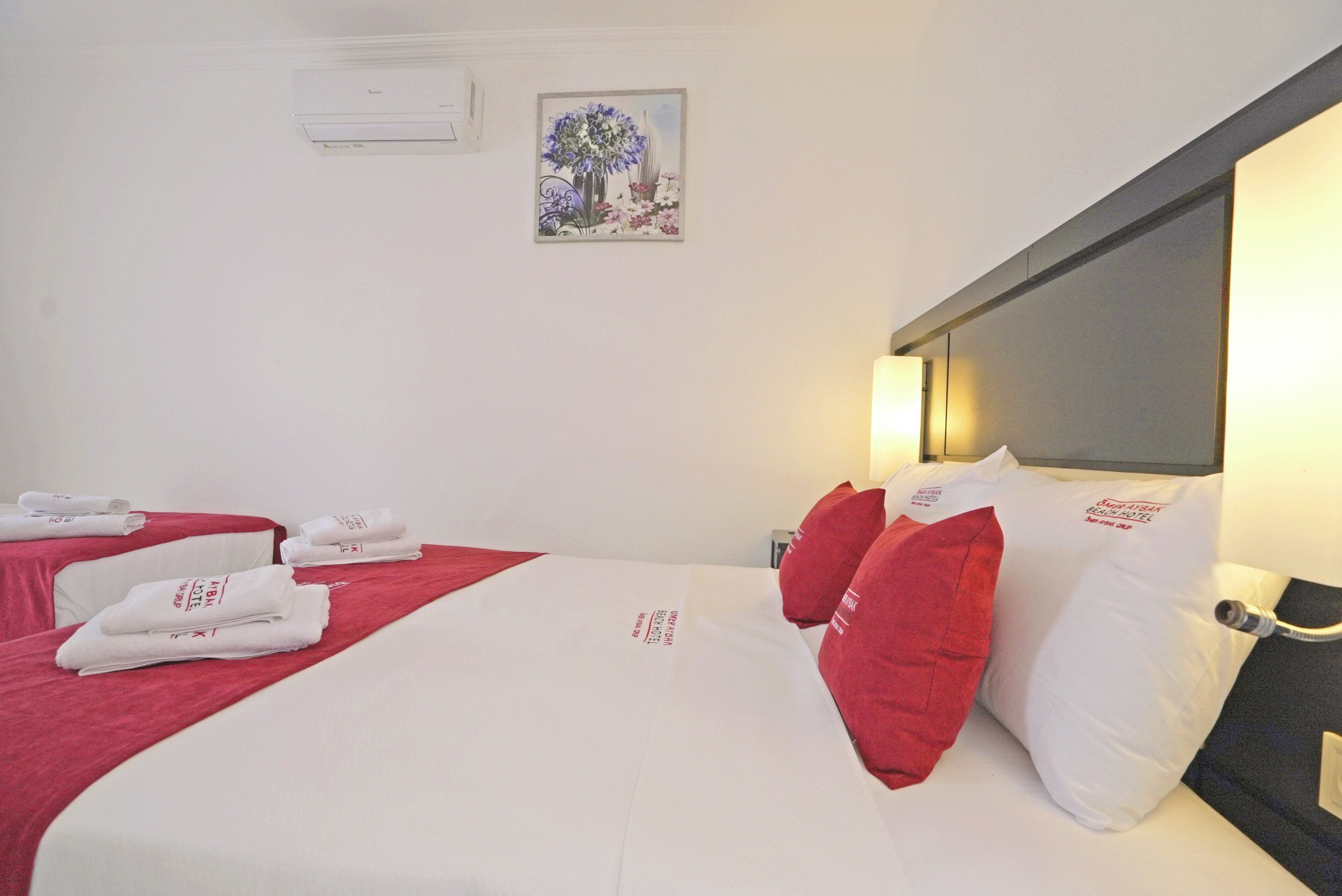 A hotel room with two beds, red and white bedding, a painting on the wall, and a bedside table with a lamp.