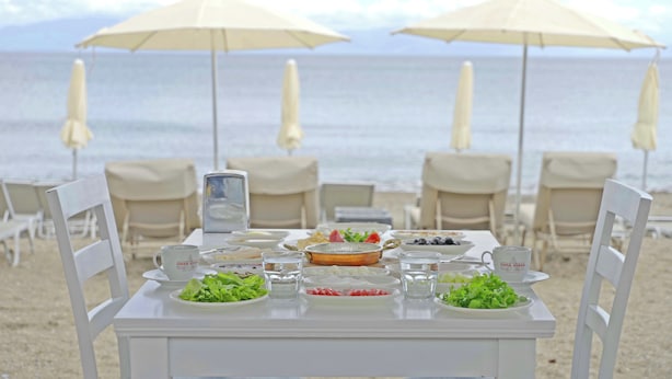 A beachside table set with plates, glasses, and a bowl of salad, surrounded by beach chairs and umbrellas. - Ömer Aybak Beach Hotel (Bodrum)