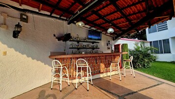 Un área de bar al aire libre con una barra de ladrillo, sillas de metal blanco y un televisor montado en la pared.
