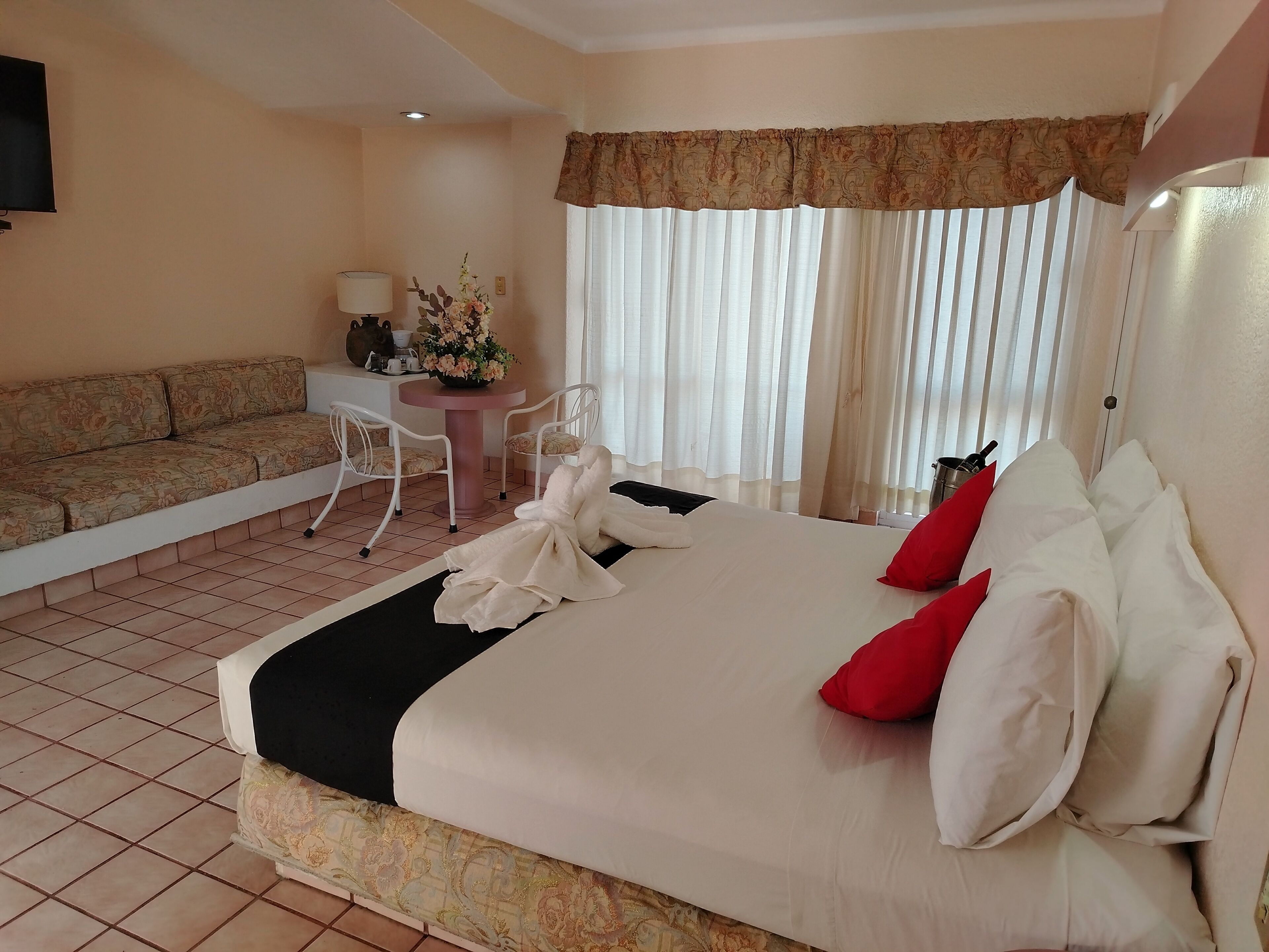 A hotel room with a bed, a sofa, a small table with a lamp, and a window with curtains.