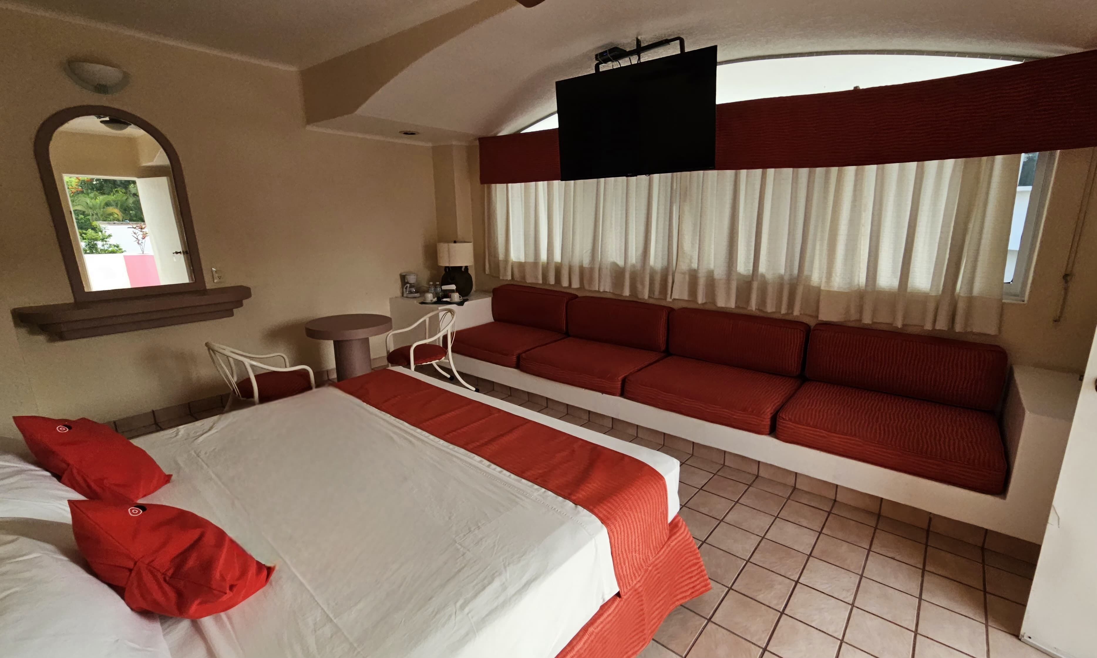 A hotel room with a large bed, a sofa bed, a small table with a chair, a TV mounted on the wall, and a window with curtains.
