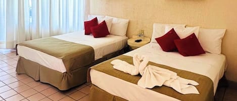 A hotel room with two beds, each with a brown skirted base, white linens, and red pillows. There is a small bedside table with a lamp, and the room has a tiled floor.