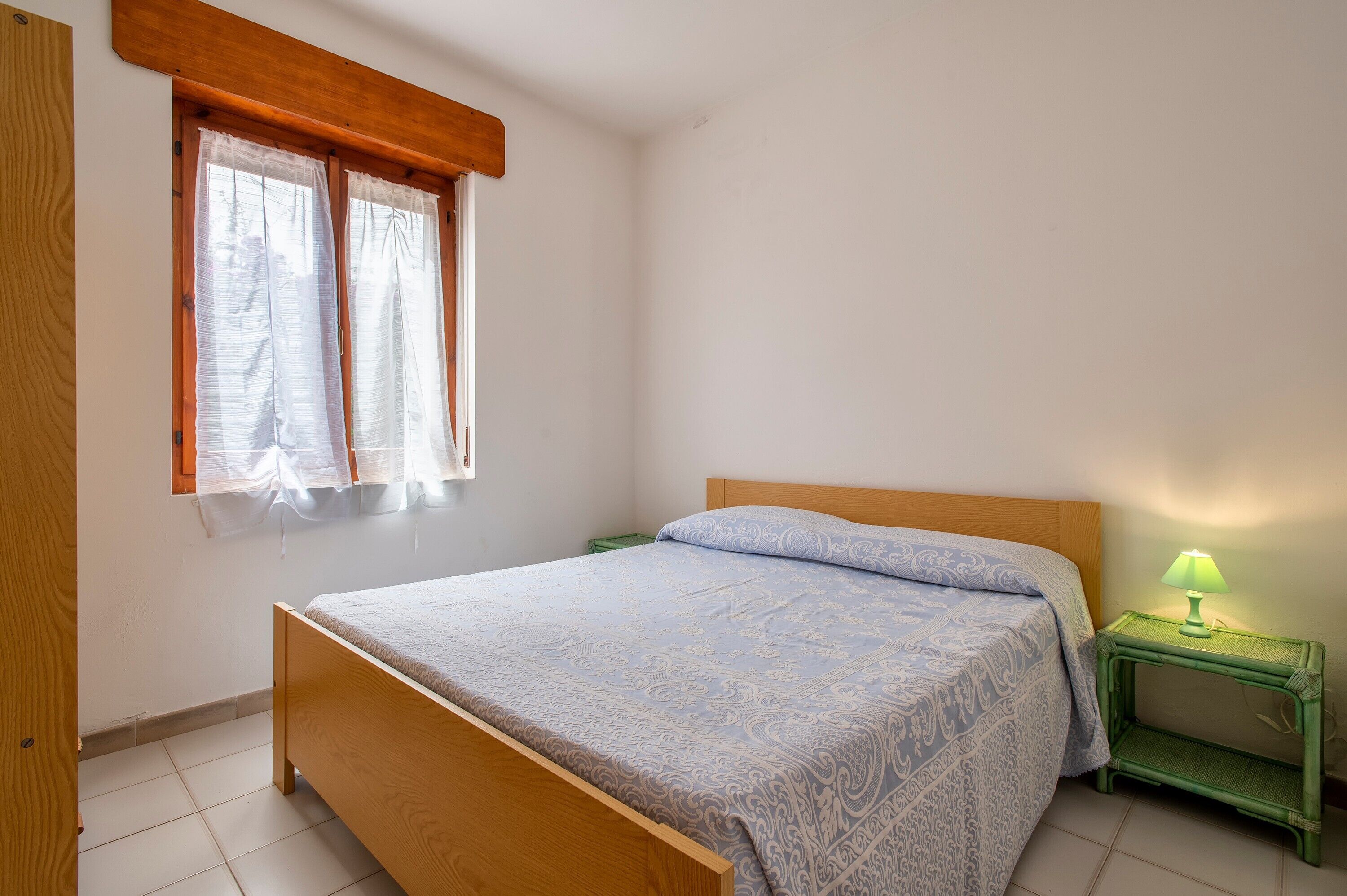 Un dormitorio con cama de madera, una mesita de noche verde con lámpara y una ventana con cortinas.