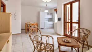 Una cocina con gabinetes blancos, un comedor con sillas de mimbre y vistas a través de una ventana.