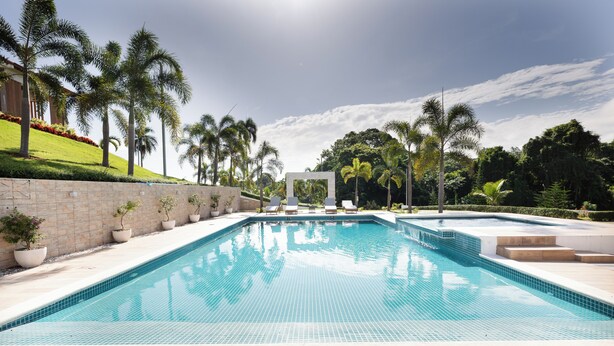 A swimming pool with tiled edges, surrounded by palm trees and a stone wall. - Hermosa villa rodeada de montañas, Combina elegancia moderna con la naturaleza. (Bonao)