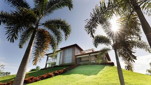 A modern house with a sloped roof, surrounded by lush green grass and palm trees. - Hermosa villa rodeada de montañas, Combina elegancia moderna con la naturaleza. (Bonao)