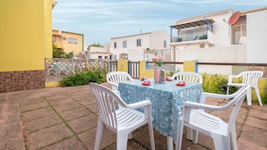 Une terrasse avec une table dressée pour deux personnes, entourée de chaises en plastique blanches, d’une nappe à motifs et d’un vase rempli de fleurs.