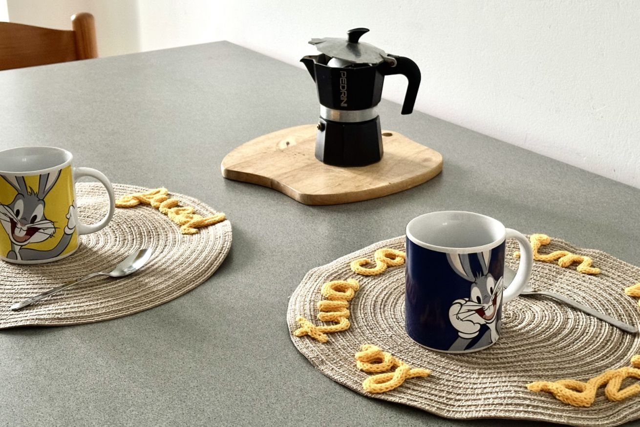 Two coffee cups on placemats with cartoon characters, a coffee maker on a wooden stand, and a woven placemat on a table.