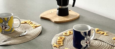 Two coffee cups on placemats with cartoon characters, a coffee maker on a wooden stand, and a woven placemat on a table.