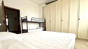 Una cama individual con colchón blanco y una cama litera a la izquierda. Se observa un armario con varias puertas y una puerta que da a otra habitación.