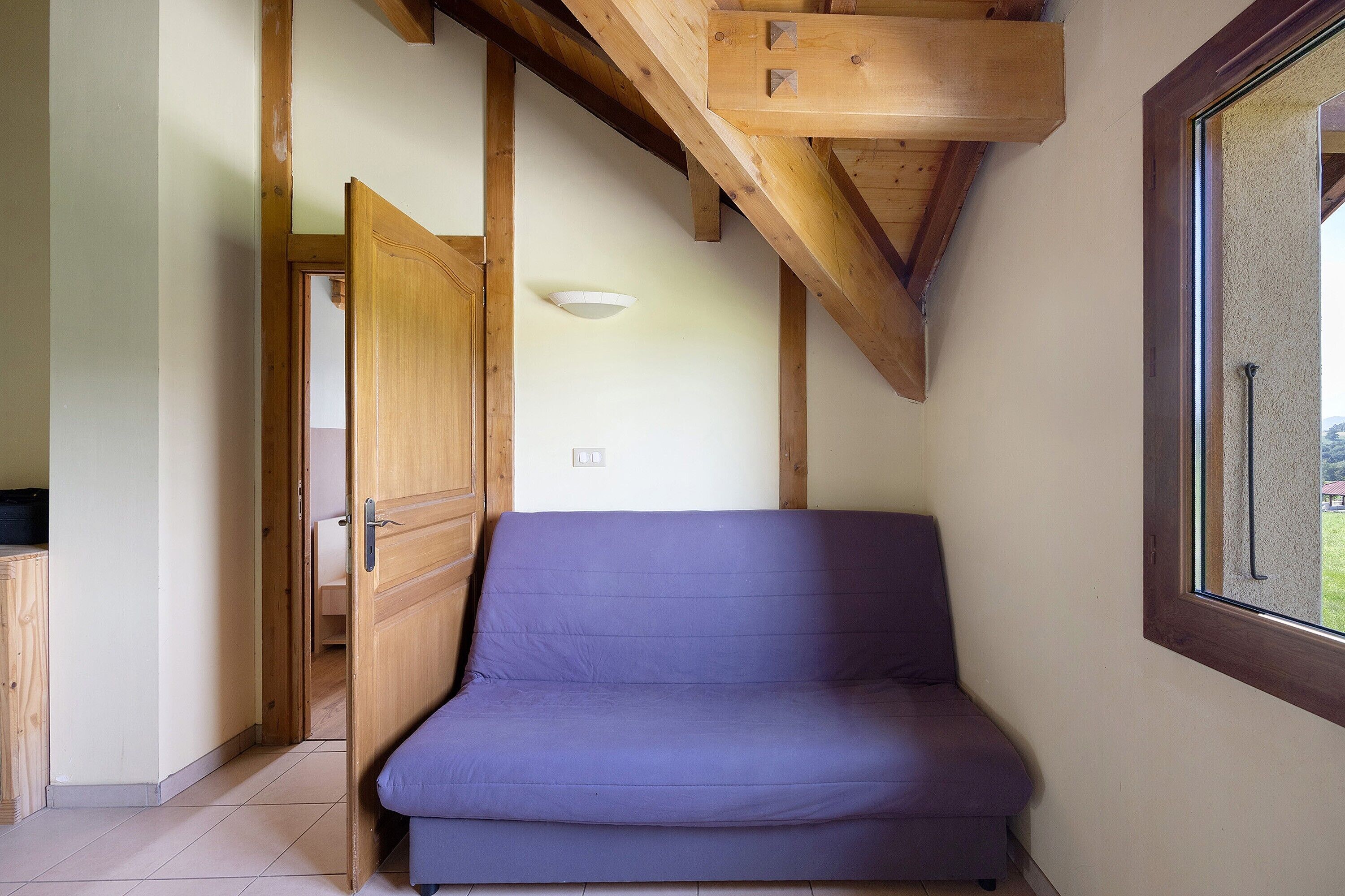 Une pièce avec un plafond en bois, un matelas violet, une porte et une fenêtre.