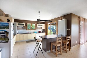 A modern kitchen with a central island, wooden chairs, and a view of the outdoors through large windows. - Cottage 'Villa De St Off' with Mountain View, Private Terrace and Wi-Fi (Saint-Offenge)