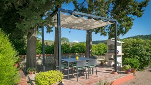 Un patio techado con una mesa y sillas, rodeado de plantas en macetas y una parrilla.