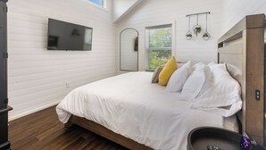 A bedroom with a large bed, a flat-screen TV mounted on the wall, a window with blinds, and a ceiling fan.
