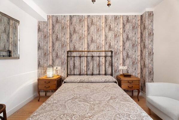 Quarto com cama, duas mesas de cabeceira, uma cadeira branca e um painel de cama de madeira.