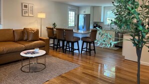 A living room with a brown leather sofa, a small round coffee table, and a kitchen area with a refrigerator and bar stools. - Brand New Four Bedroom Home Near Beach and Town (Chatham)
