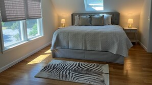 A bedroom with a bed, two bedside tables with lamps, a zebra-patterned rug, a window with blinds, and a skylight. - Brand New Four Bedroom Home Near Beach and Town (Chatham)