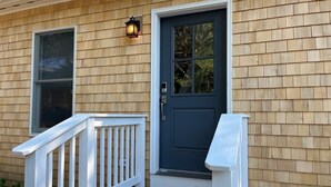 A house with a dark blue front door, a white front porch railing, and a window with a view of trees. - Brand New Four Bedroom Home Near Beach and Town (Chatham)