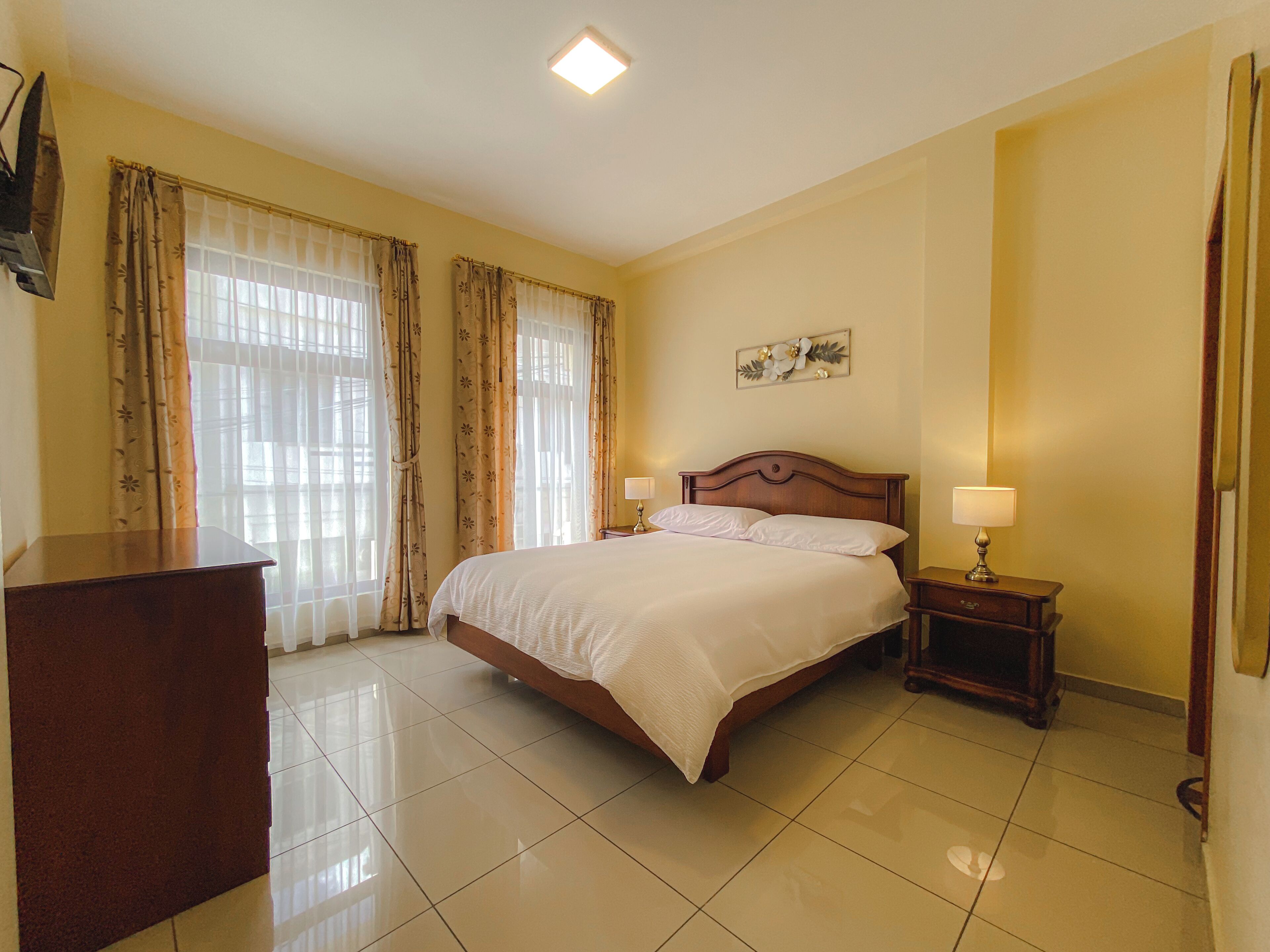 Habitación de hotel con una cama grande, una mesita de noche con lámpara, un aparador y una ventana con cortinas.