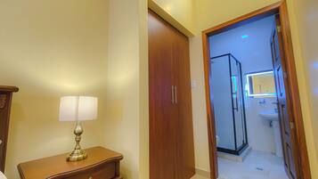 Corredor de hotel com porta de madeira, mesa de cabeceira com abajur e banheiro visível através de uma porta aberta.