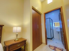 Corredor de hotel com porta de madeira, mesa de cabeceira com abajur e banheiro visível através de uma porta aberta.