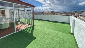 Une terrasse sur le toit avec un revêtement d’herbe artificielle, un espace clos en verre avec des sièges, et une vue sur le paysage urbain.