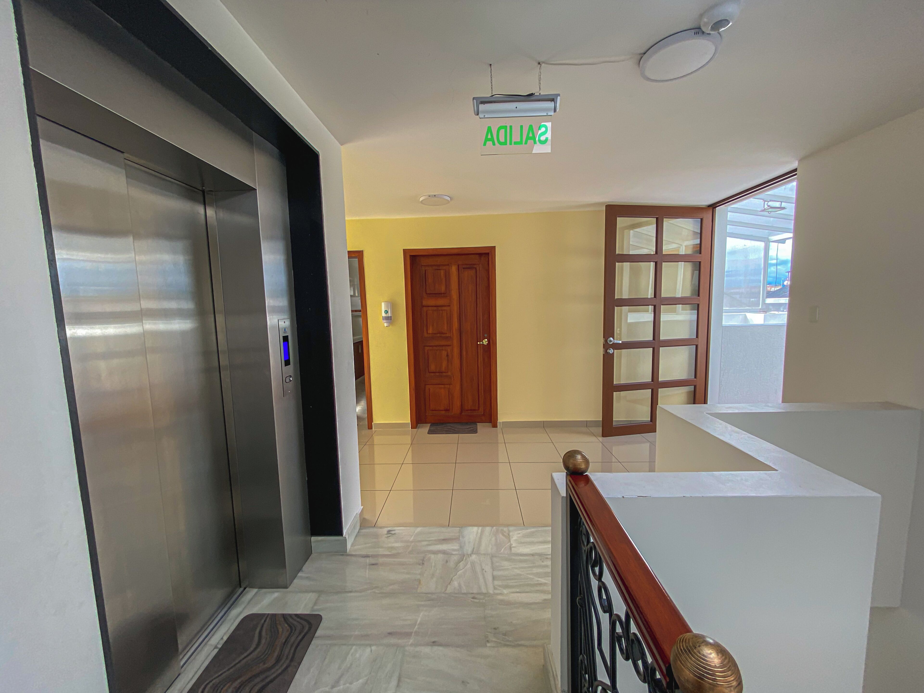 A hallway with a modern elevator, a wooden door, and a view of the outside through a window.