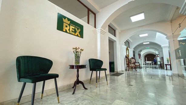 Un hall d’hôtel avec un long couloir, des fauteuils à revêtement vert, un vase rempli de fleurs et une enseigne indiquant « GRAN HOTEL REX ».