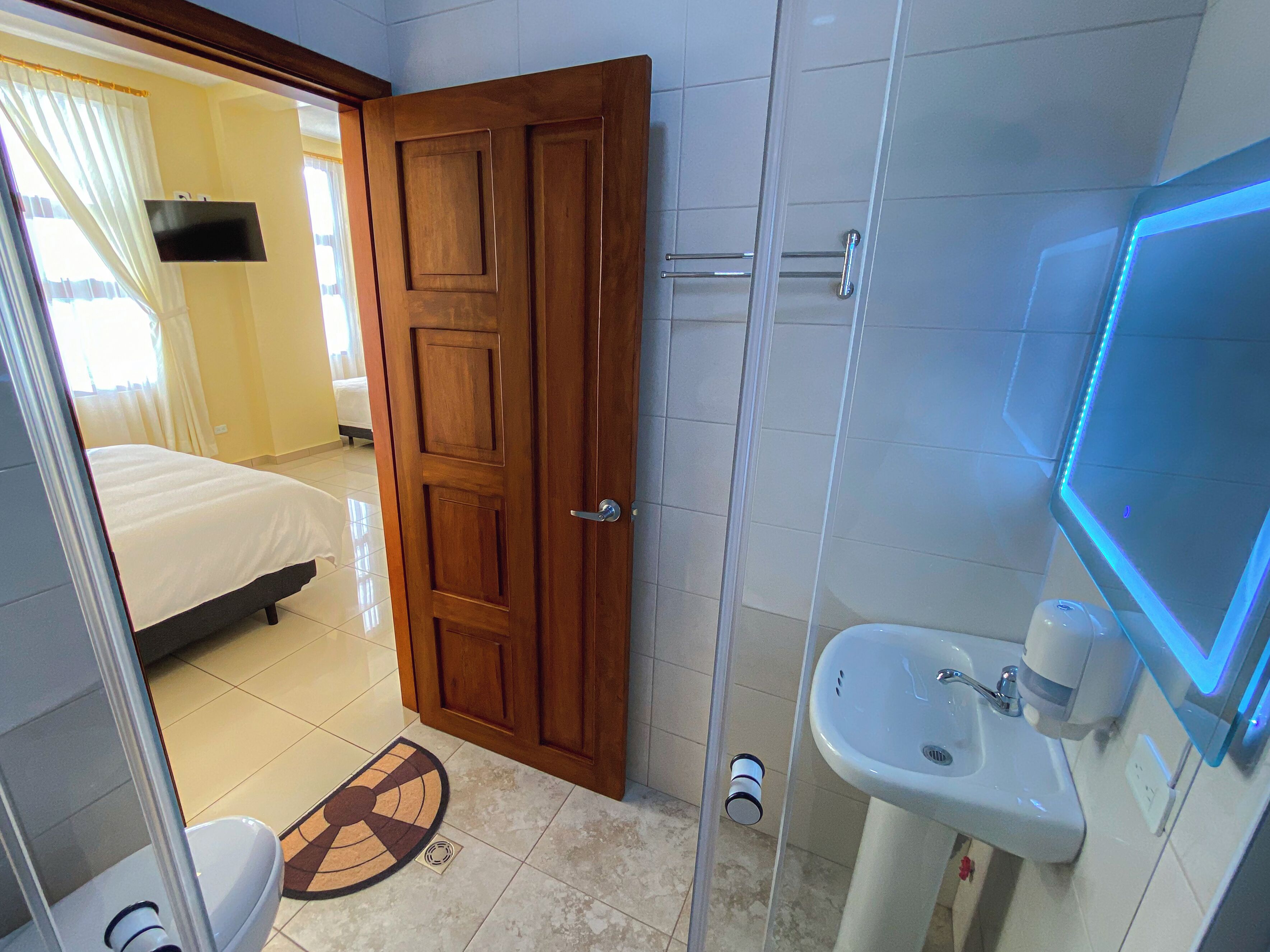Une chambre d’hôtel avec une porte en bois, un lit, une salle de bain avec un lavabo et un miroir, et un espace douche.