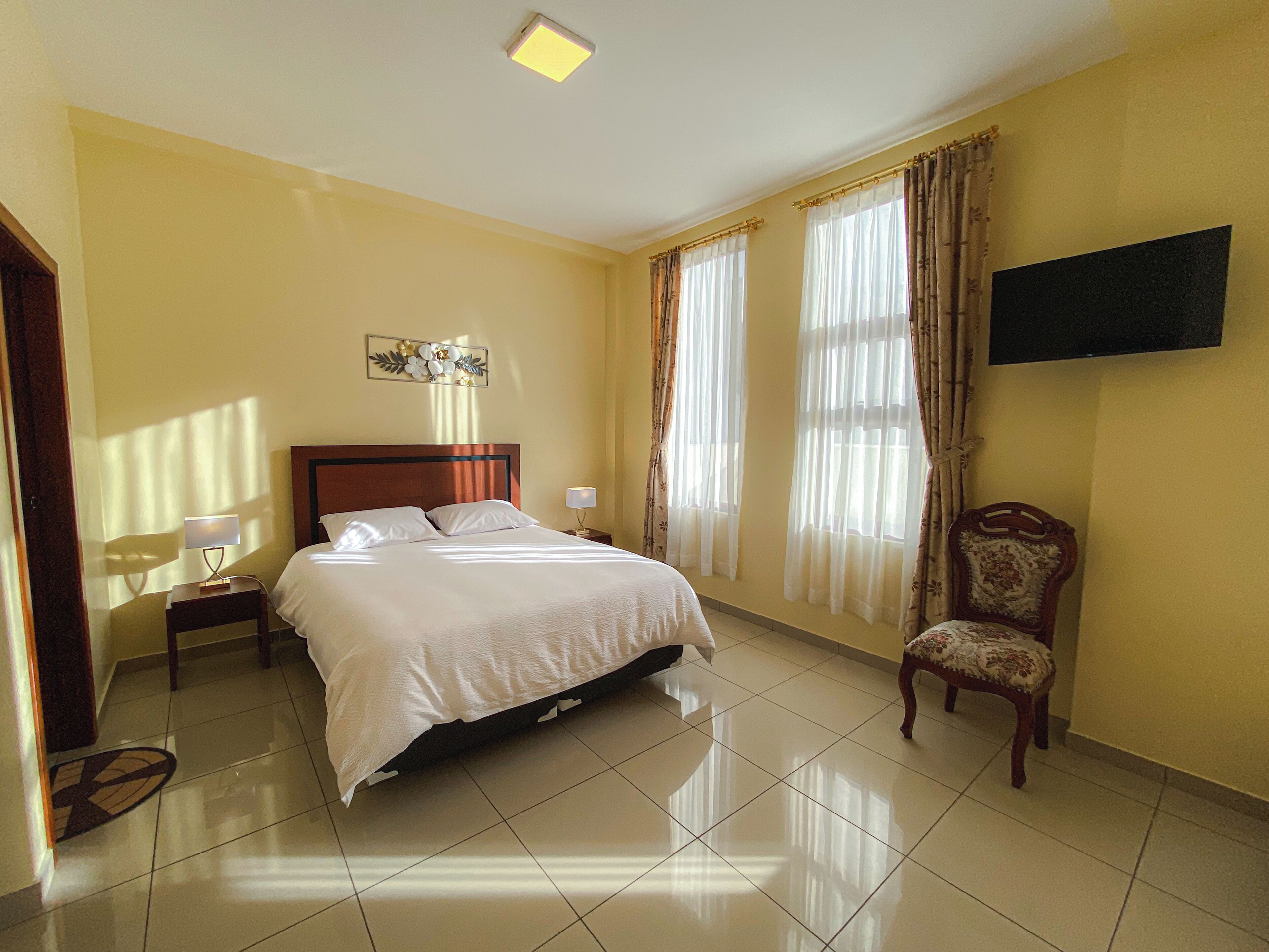 Una habitación de hotel con cama, una silla, televisor montado en la pared y ventana con cortinas.