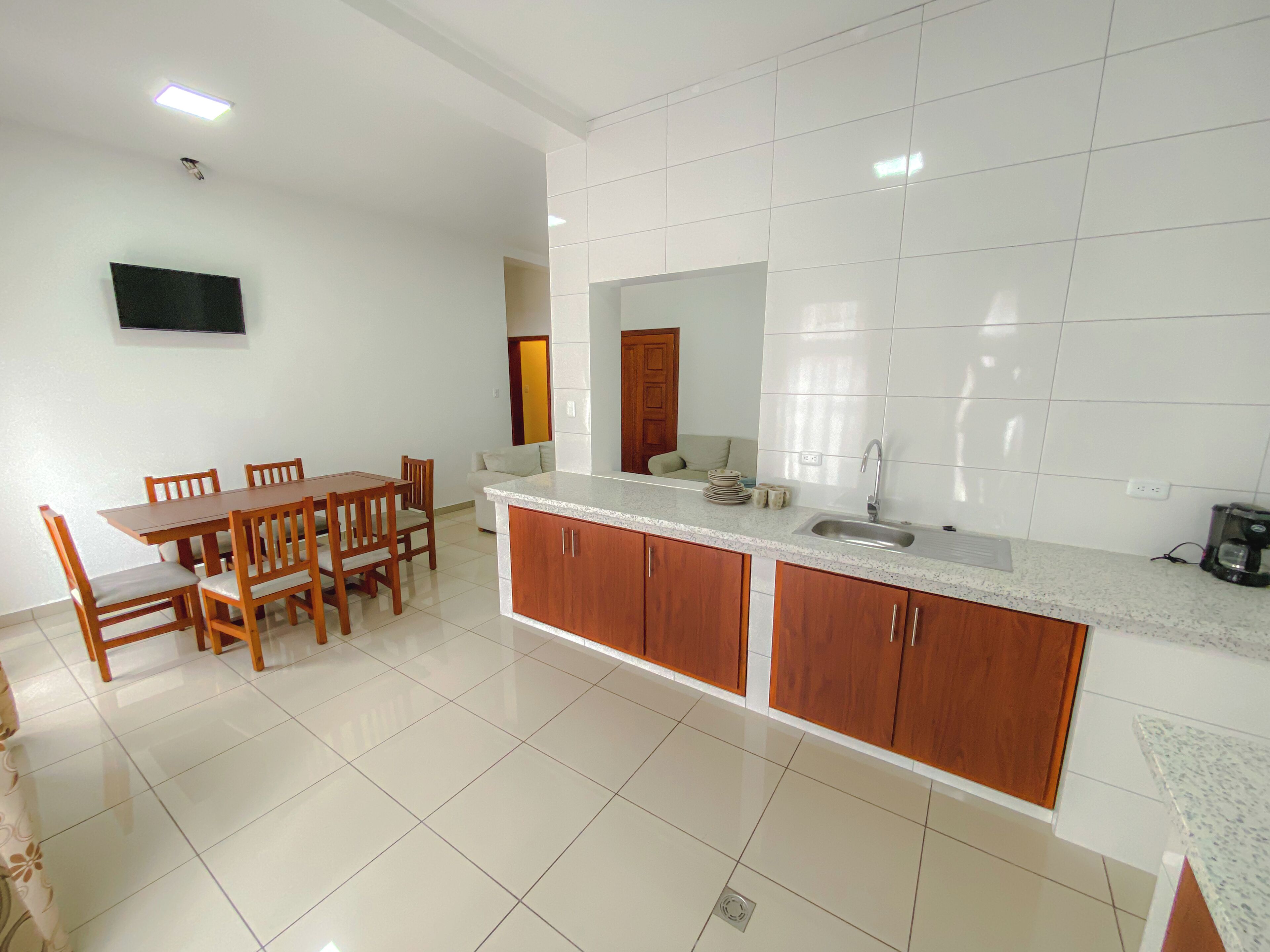 A modern kitchen with white tiles, wooden cabinets, a granite countertop, and a dining area with a table and chairs.