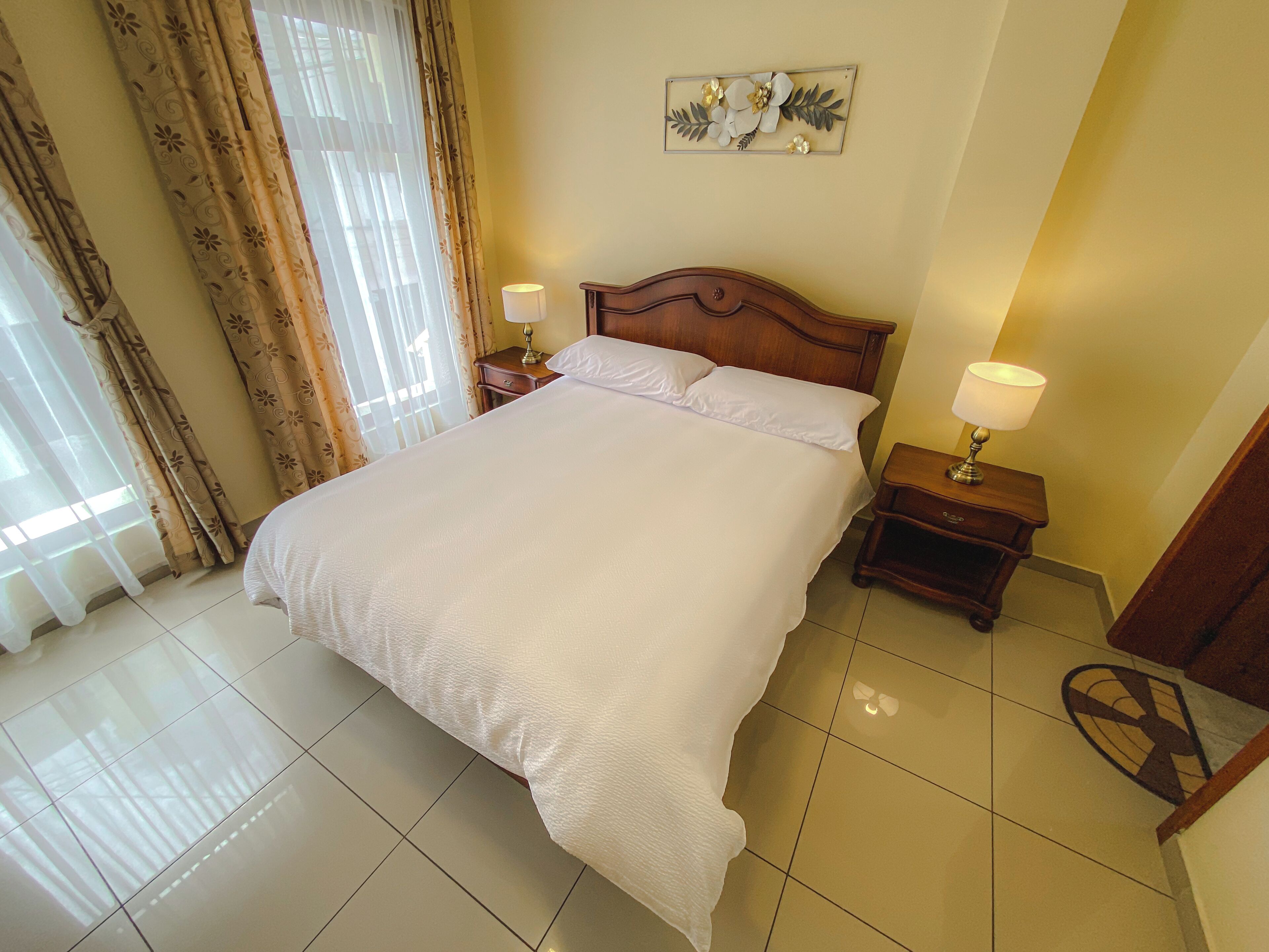 A neatly made bed with white linens, two bedside tables with lamps, a decorative wall piece, and a window with curtains.