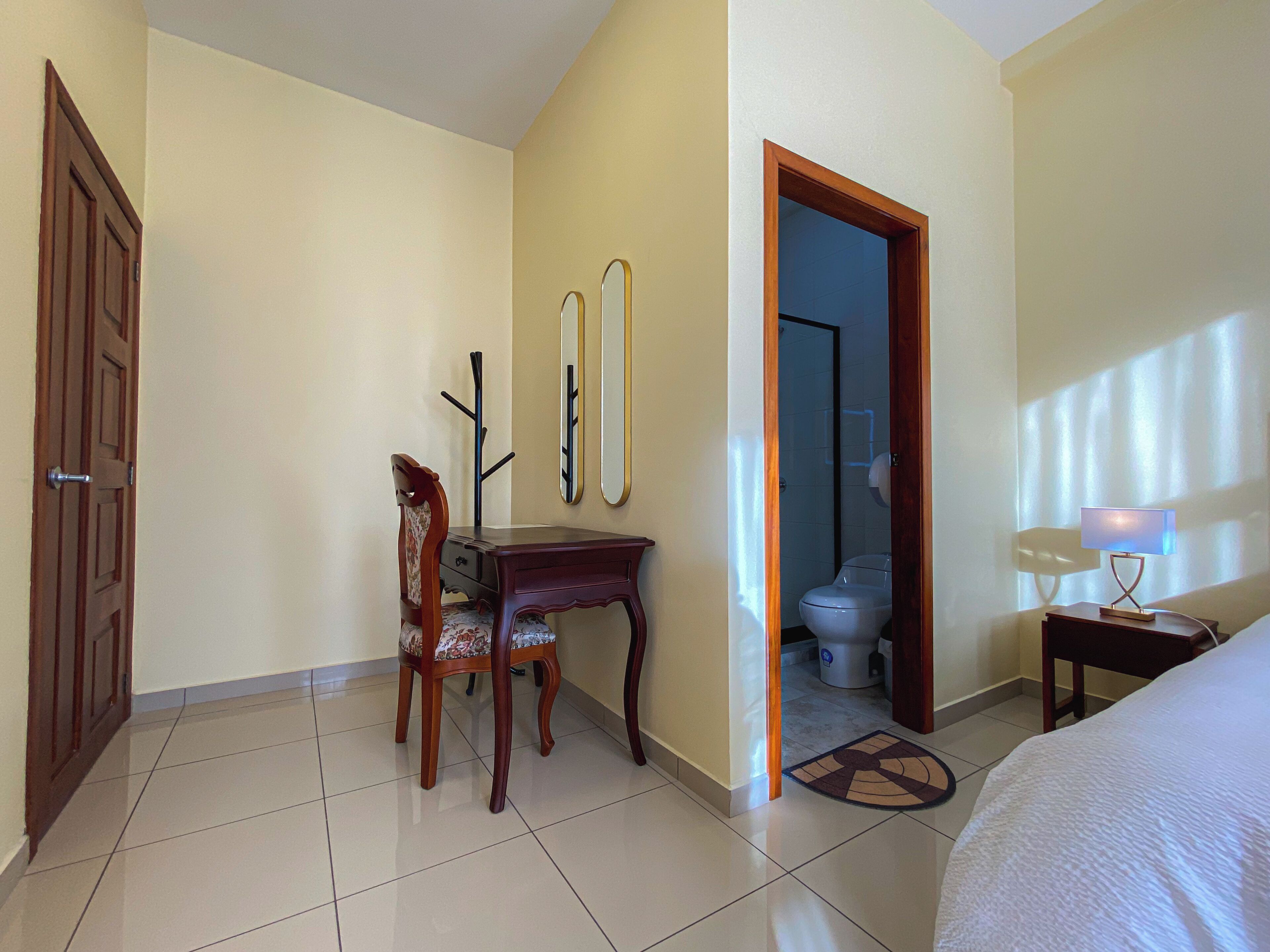 A hotel room with a wooden desk, a chair, a bed, a nightstand with a lamp, and a bathroom visible through an open door.