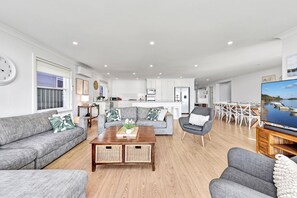 Ein modernes Wohnzimmer mit Sofa, Sessel und Couchtisch. Ein Essbereich mit Tisch und Stühlen ist ebenfalls vorhanden. Im Hintergrund ist eine Küche zu sehen.