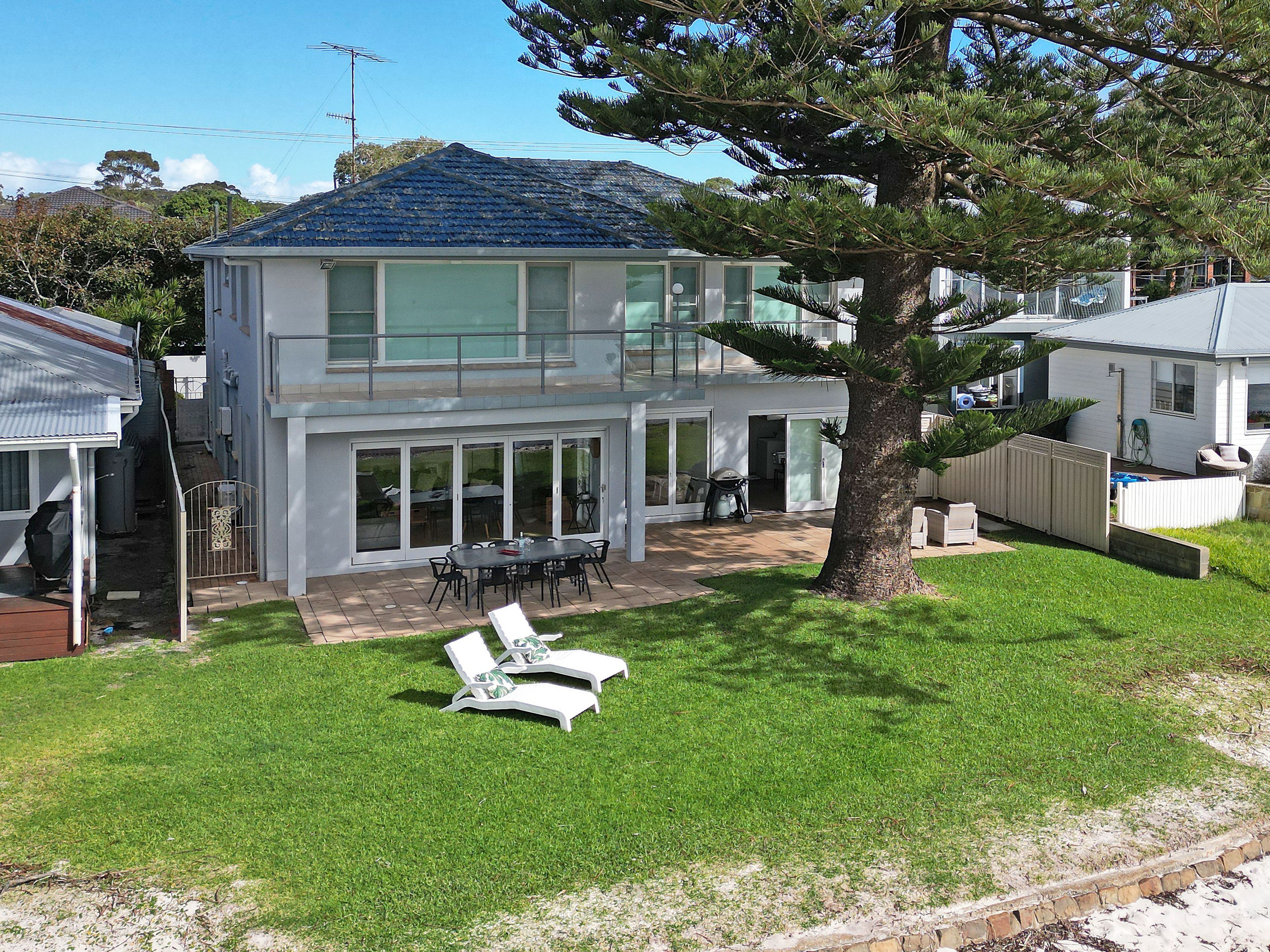 A two-story house with a patio, a large tree, and a lawn with two lounge chairs.