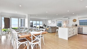 A spacious living area with a dining table set for six, a kitchen with modern appliances, and large windows offering a view of the sea. - Filoli - A breathtaking waterfront (Salamander Bay)