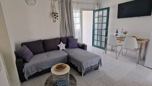 Un salón moderno con un sofá gris, una pequeña mesa redonda con un cuenco, una silla de madera, un televisor montado en la pared y una puerta con marco verde.