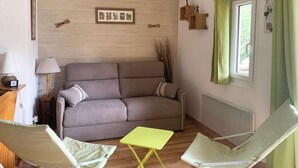 Una sala de estar acogedora con un sofá gris, dos sillas blancas, una mesita amarilla y una ventana con cortinas verdes.