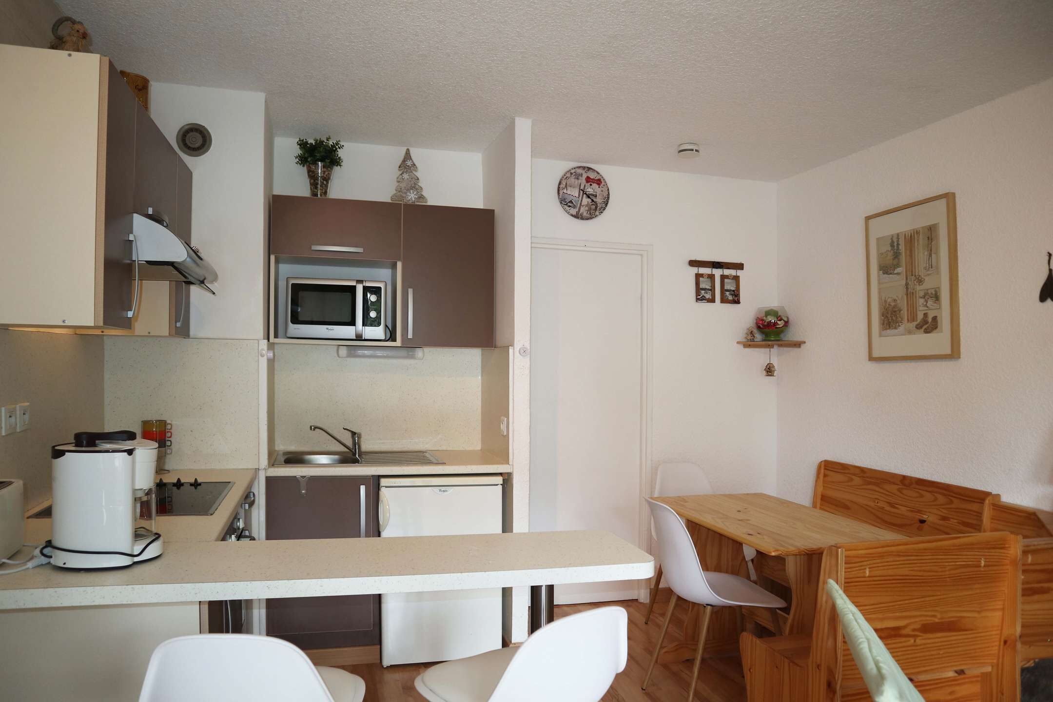 Une cuisine compacte équipée d’un four à micro-ondes, d’un évier et d’une machine à café. Un coin repas est aménagé avec une table et des chaises en bois.