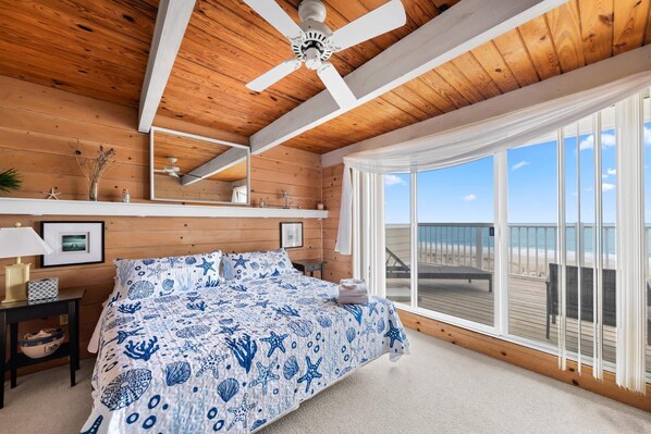 A bedroom with a large bed, wooden ceiling, a ceiling fan, a window with a view of the beach, and a nightstand with a lamp.