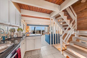 A modern kitchen with white cabinets, a black countertop, and a visible stove. There is a staircase with a wooden finish leading to a balcony with a view.